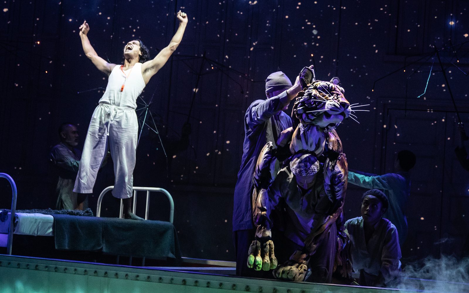Adi Dixit stands on a bed yelling with his arms in the air while Rowan Magee, Celia Mei Rubin, and Nikki Calonge